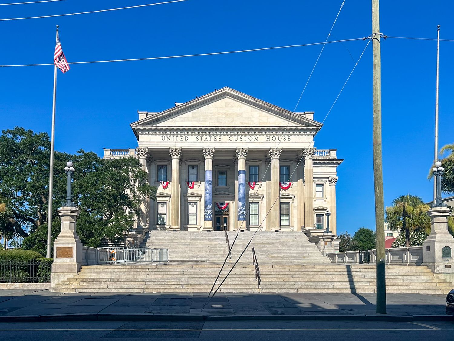 Historic neoclassical U.S. Custom House with American flag, grand steps, and decorative banners under a clear blue sky.