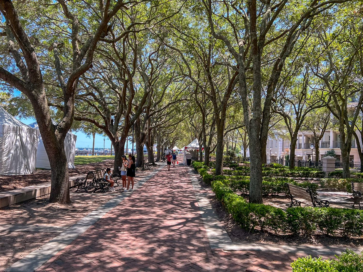 Tree-lined brick pedestrian walkway with lush greenery, benches, and people strolling on a sunny day.