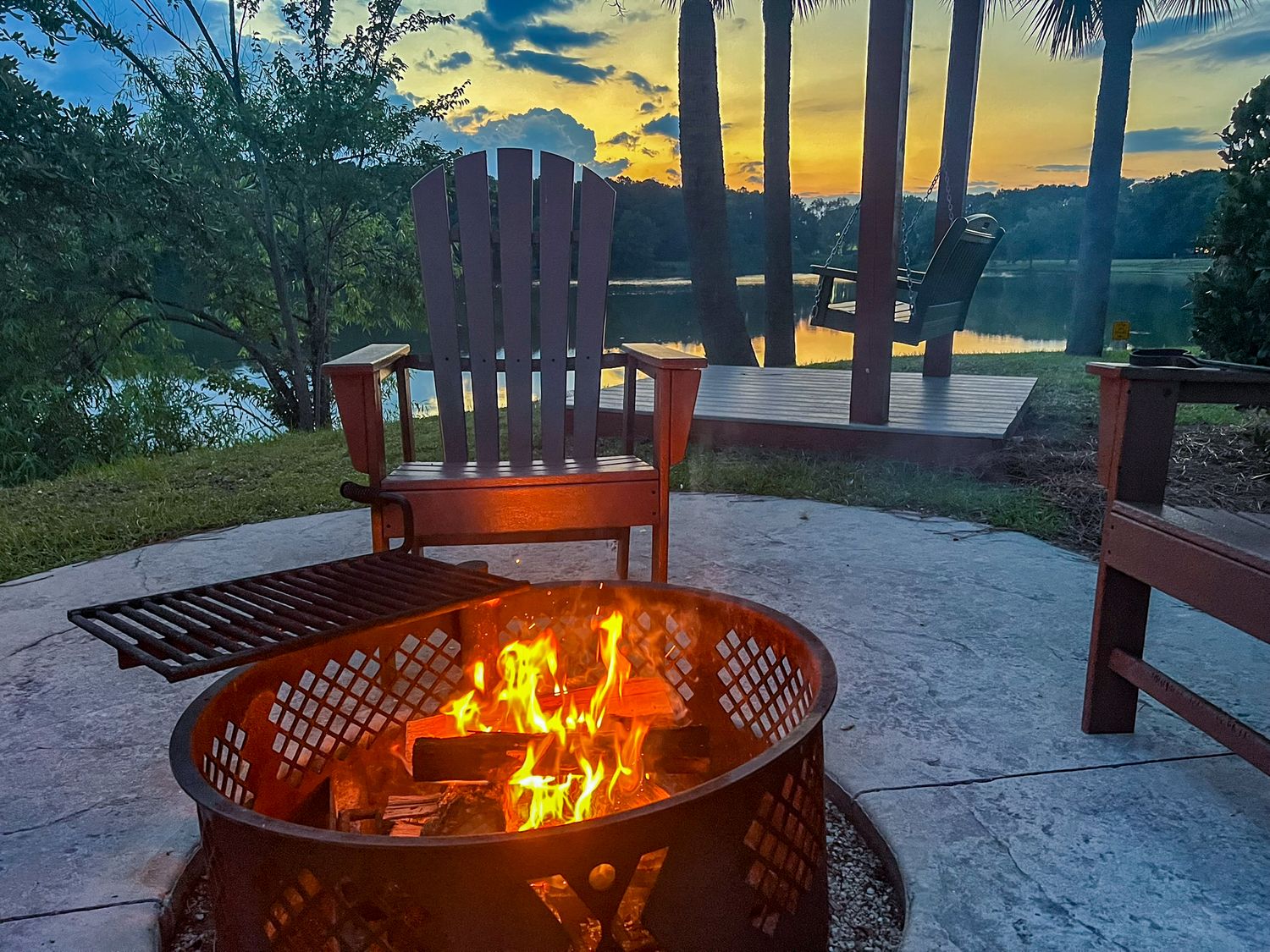 Fire pit burning at sunset by a lakeside patio with Adirondack chairs and scenic water views.