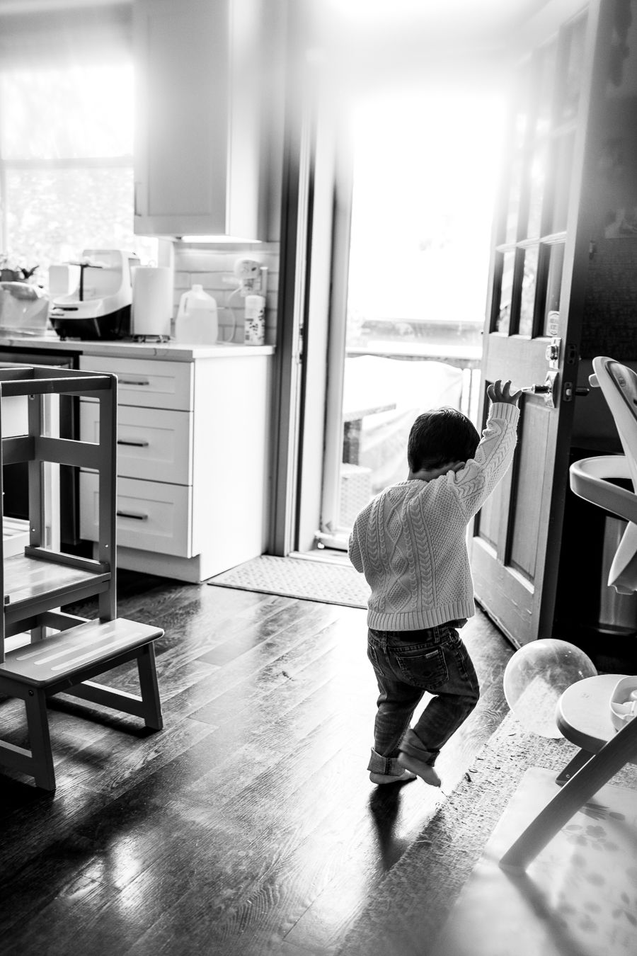 Black and white photo of a toddler reaching up to open a door in a cozy kitchen interior.