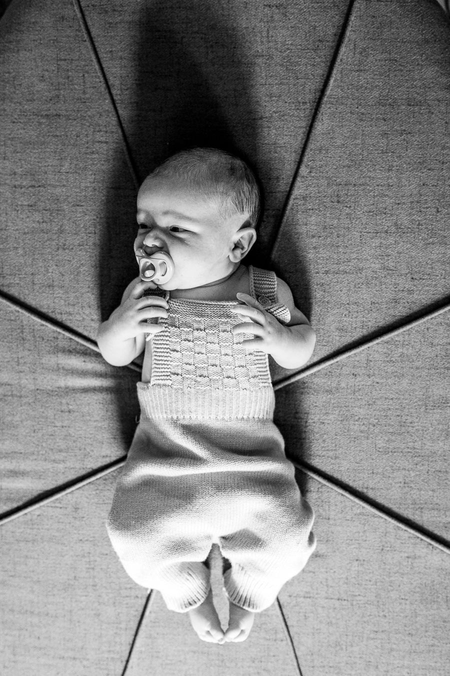 Black and white photo of a newborn baby in knitted outfit lying on cushioned surface with a pacifier.