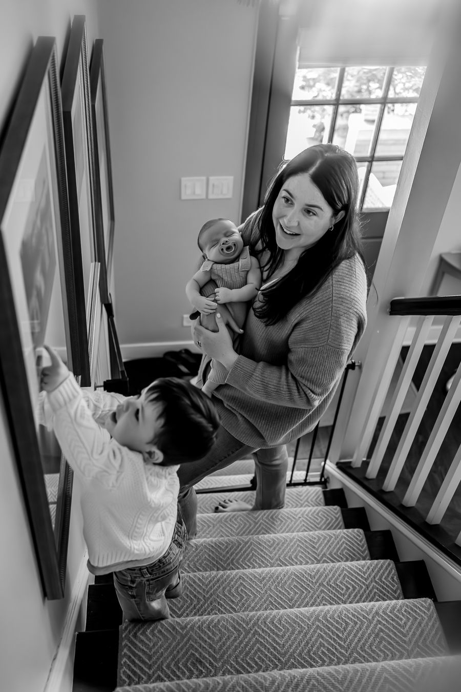 Black and white photo of a smiling mother holding a newborn on stairs with a toddler beside her.