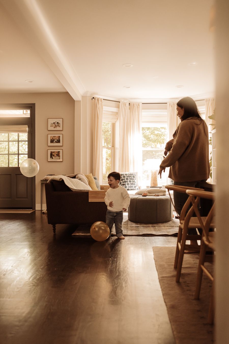 A mother watches her toddler play with balloons in a warm, sunlit living room with hardwood floors.