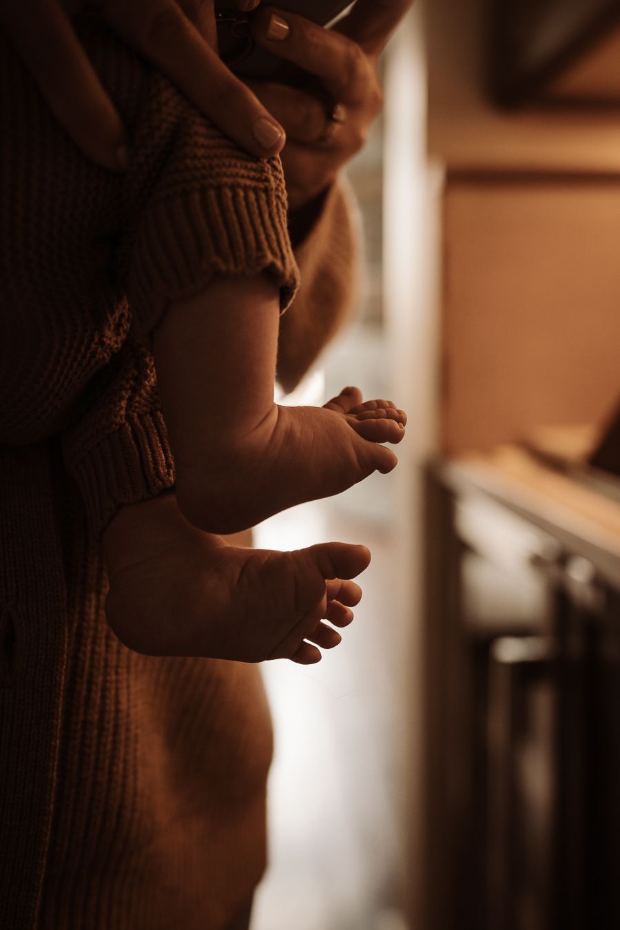 Close-up of tiny newborn baby feet dangling while being held by a parent in a warm knit sweater.