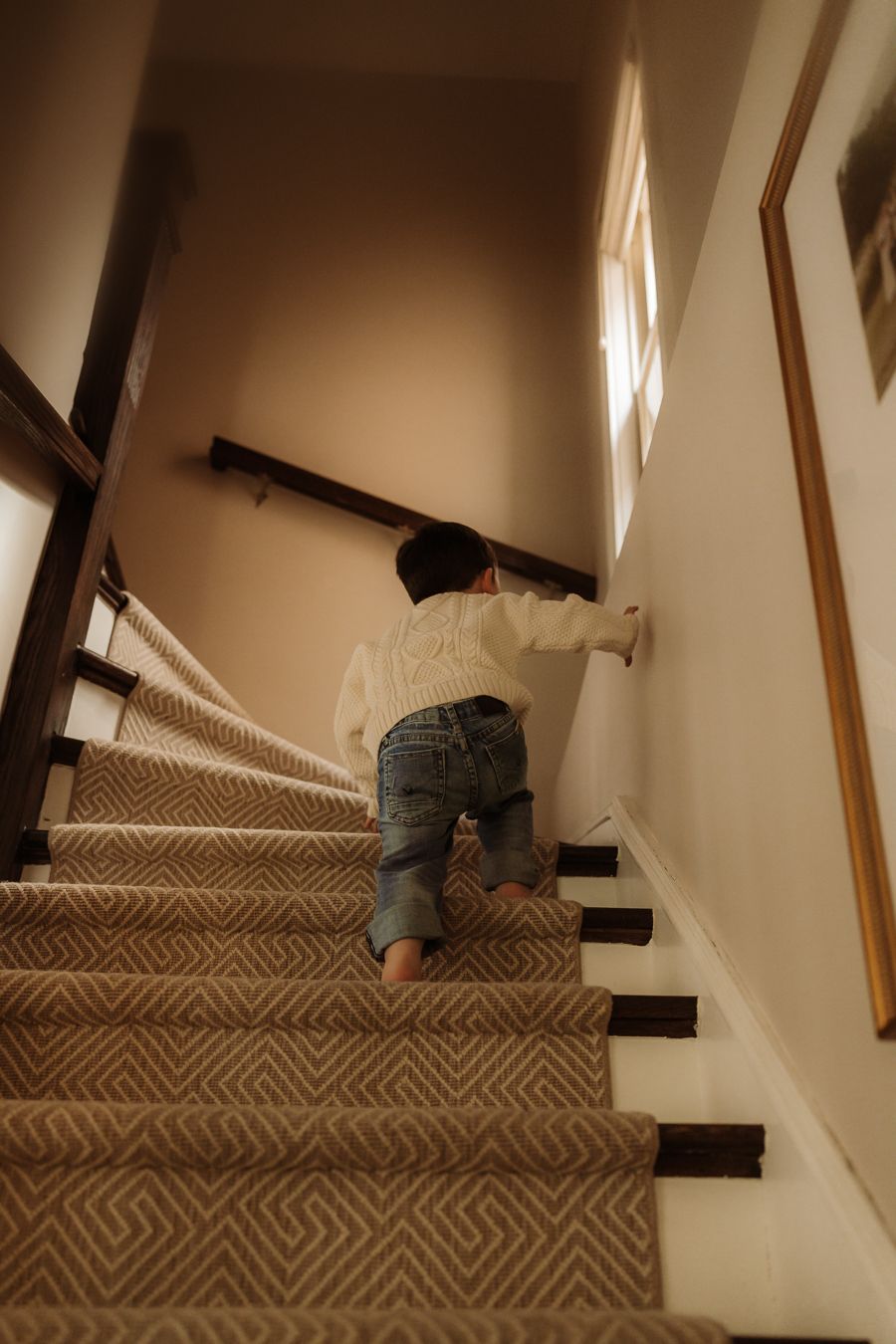 A young toddler climbs carpeted stairs indoors, wearing jeans and a white sweater, holding the railing for support.