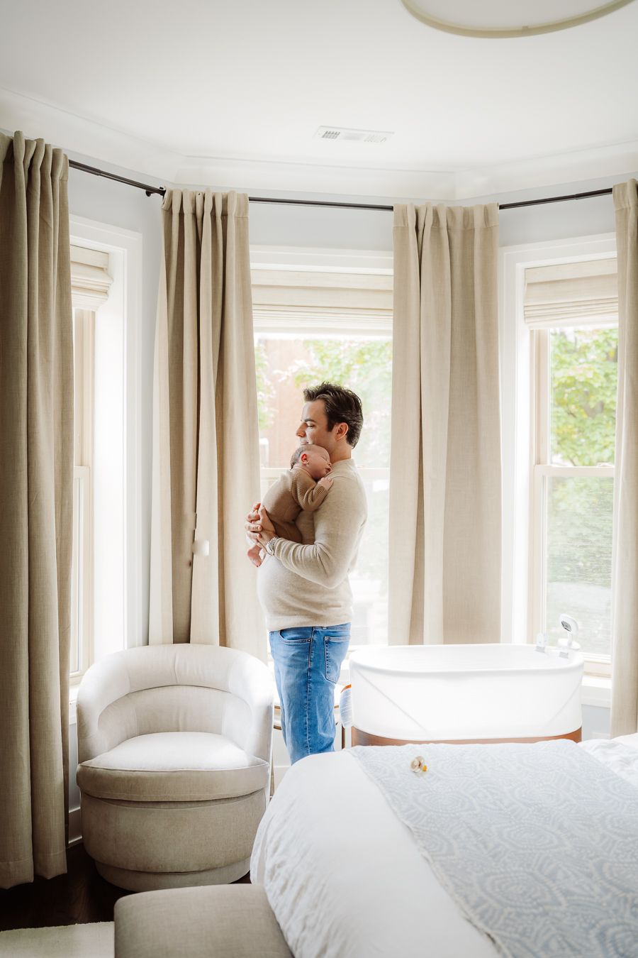 Father holding newborn baby by bay window in neutral-toned bedroom with linen curtains and white furniture.