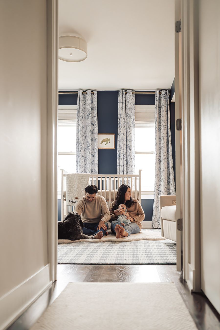 Family with baby and dog sitting on nursery floor with navy walls and floral curtains, viewed through hallway doorway.