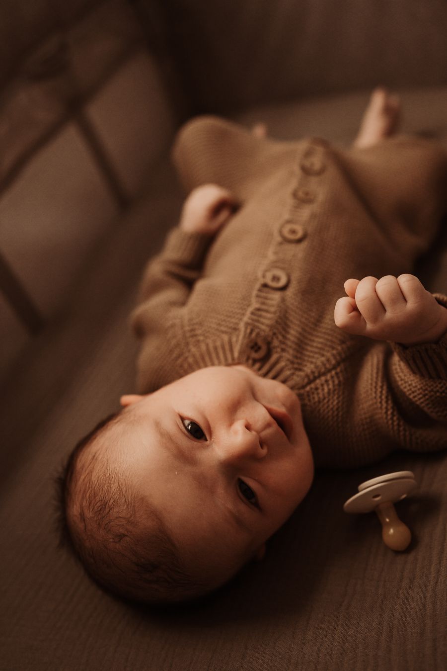Newborn baby in brown knit onesie lying in crib with pacifier nearby, looking up with dark eyes.