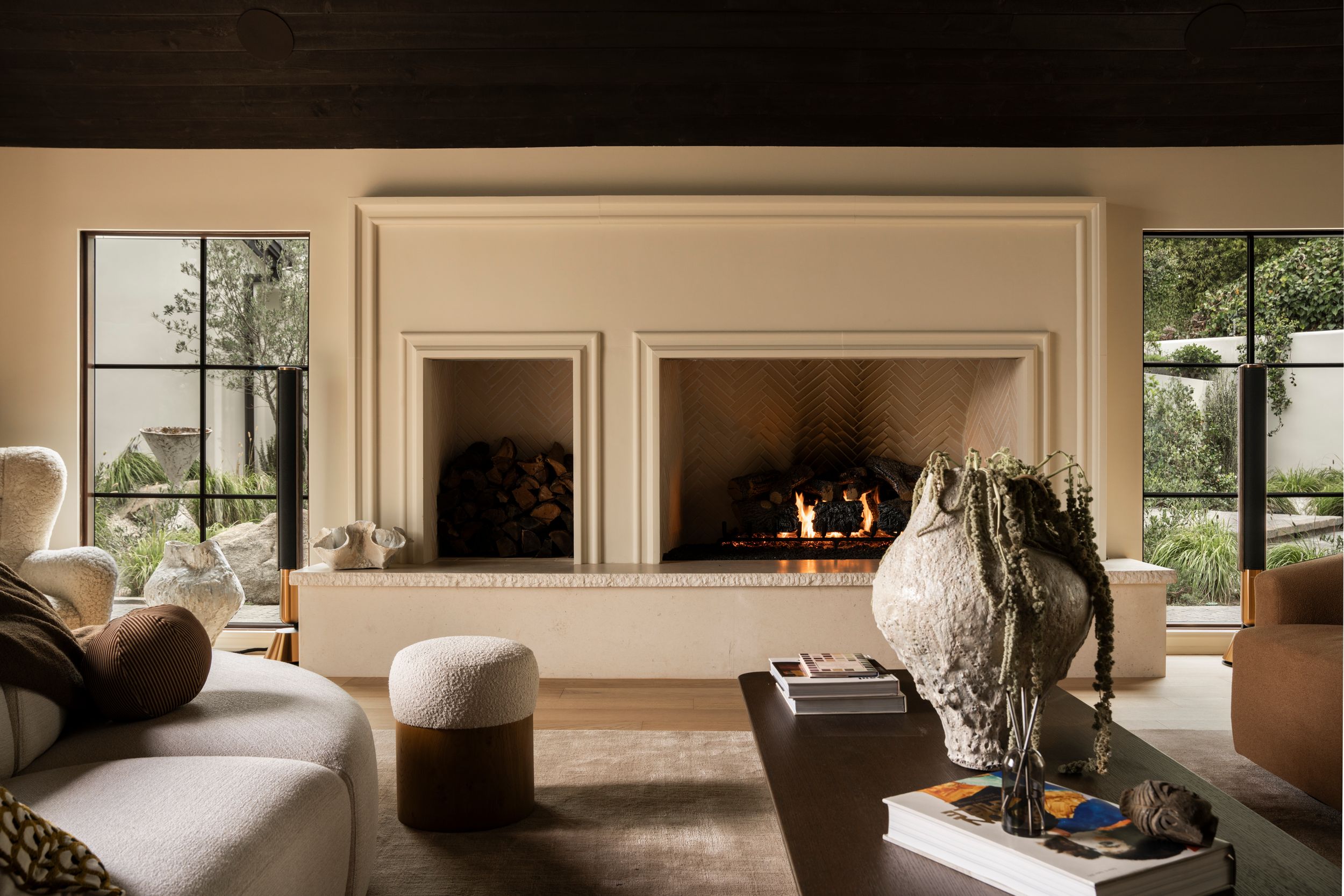 Elegant living room with a large cream fireplace, neutral furnishings, ceramic vase, and floor-to-ceiling windows overlooking greenery.