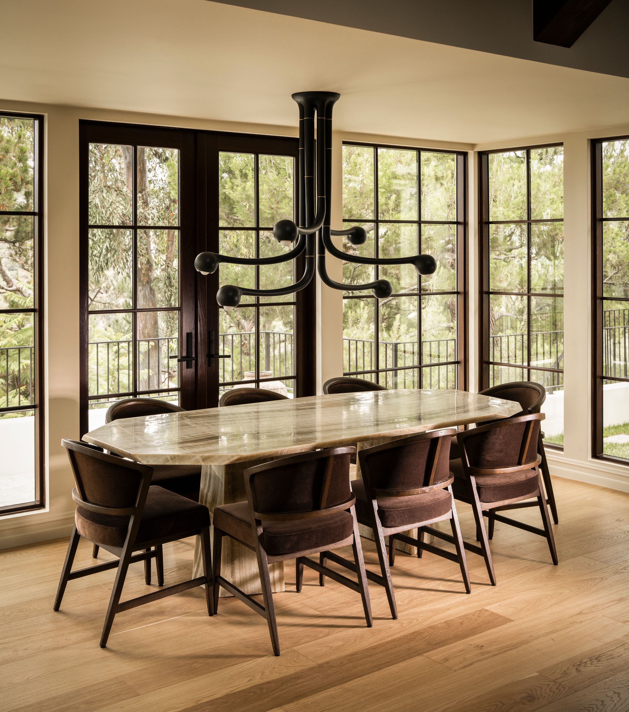 Elegant dining room with oval marble table, brown leather chairs, black chandelier, and floor-to-ceiling steel-framed windows.