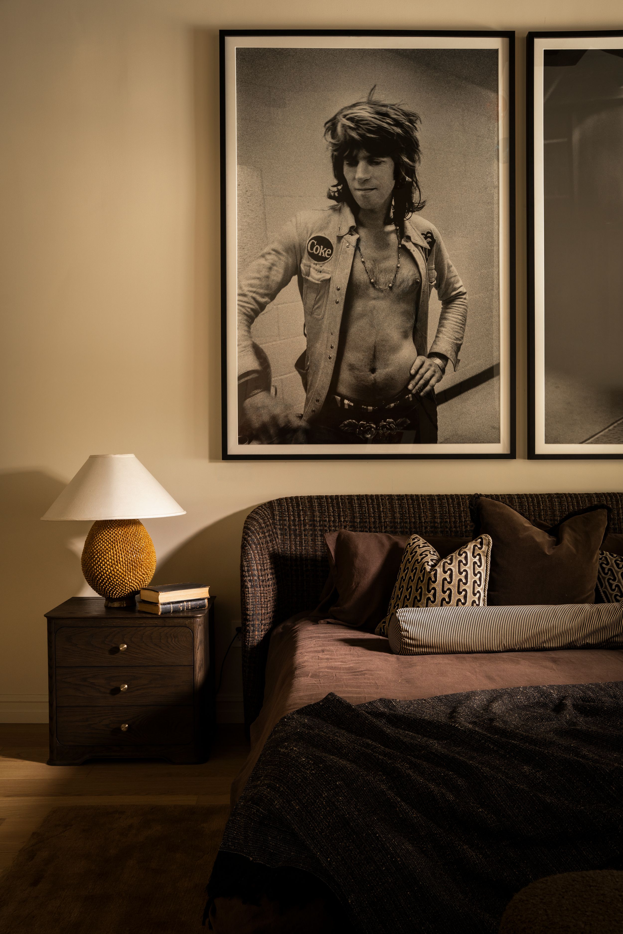 Cozy bedroom with dark bedding, wooden nightstand, lamp, and large black-and-white rock star portrait on warm beige wall.