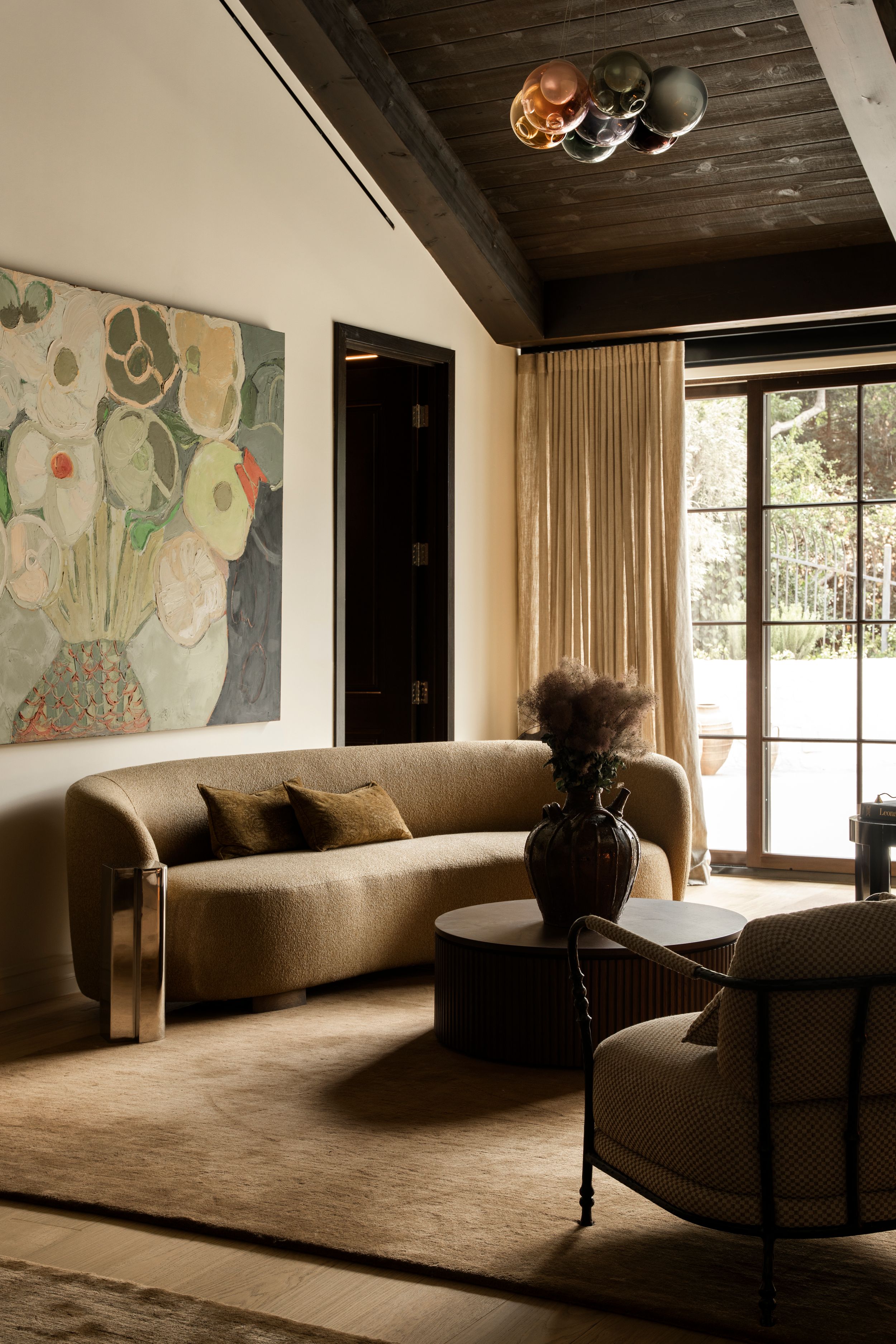 Modern living room with a curved beige sofa, floral wall art, dark ceiling, and large windows.