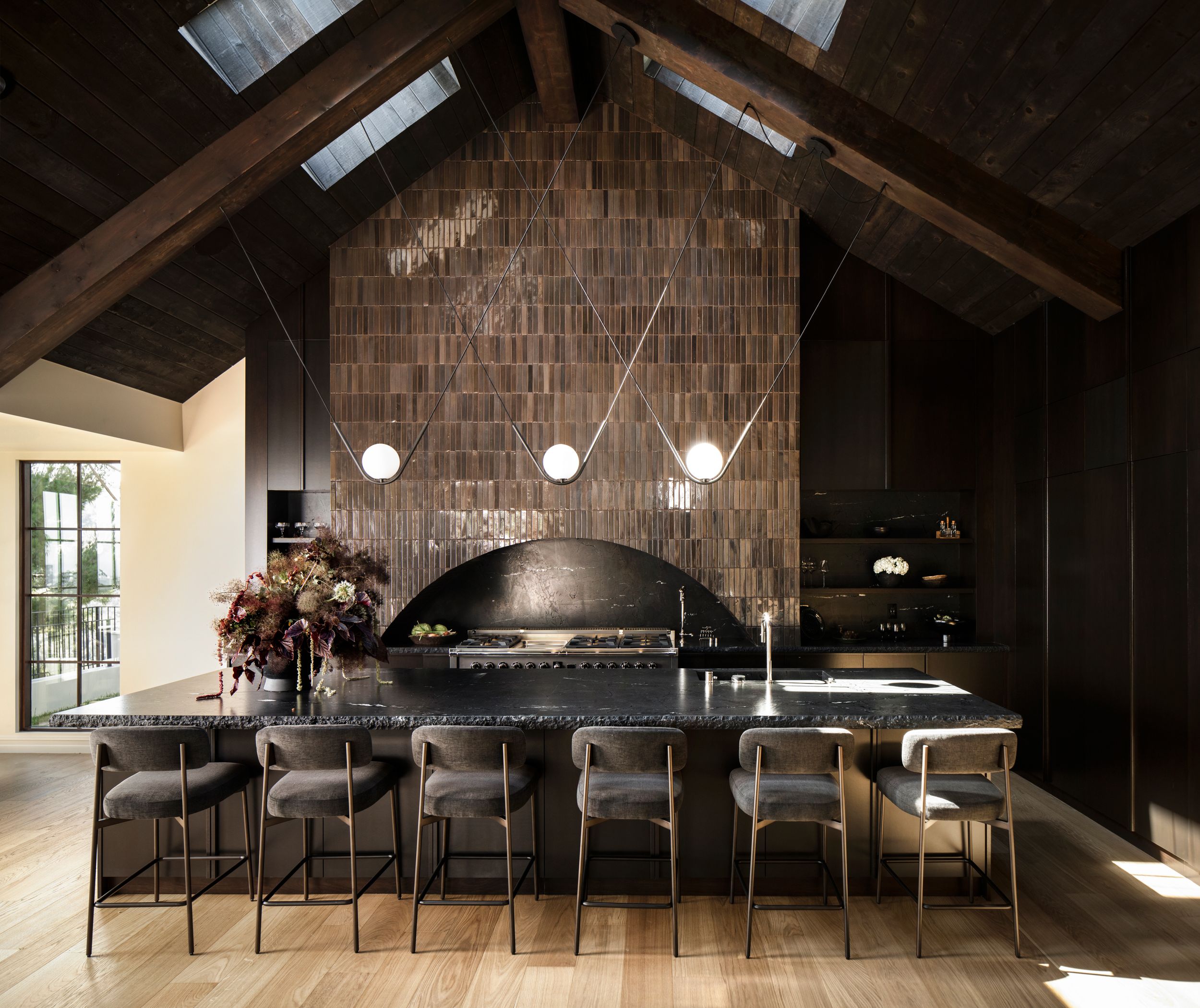 Dark modern kitchen with vaulted wood ceiling, brick arch, black island, bar stools, and pendant lights.