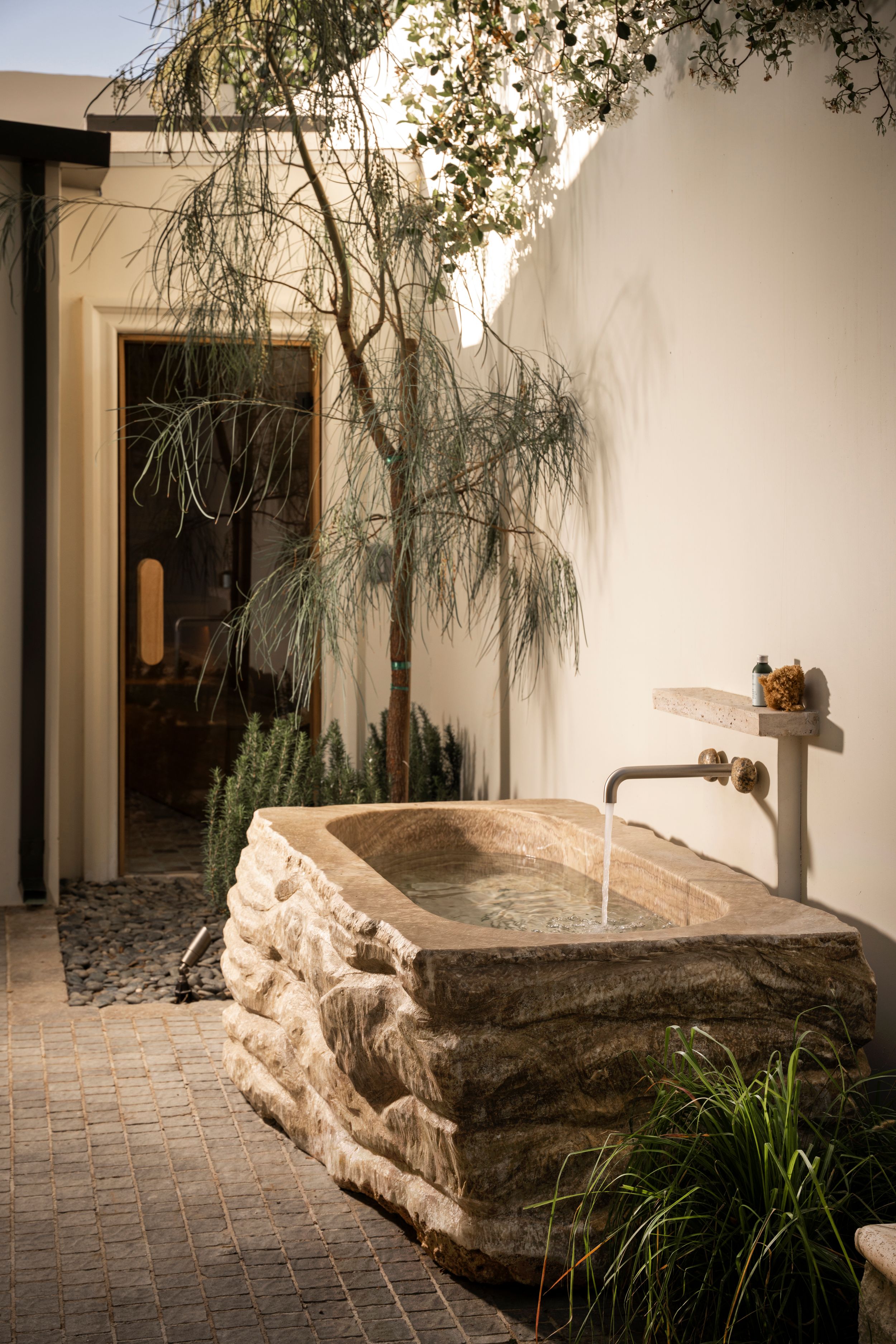 Rustic stone bathtub with flowing water faucet in an outdoor courtyard with lush greenery and white walls.