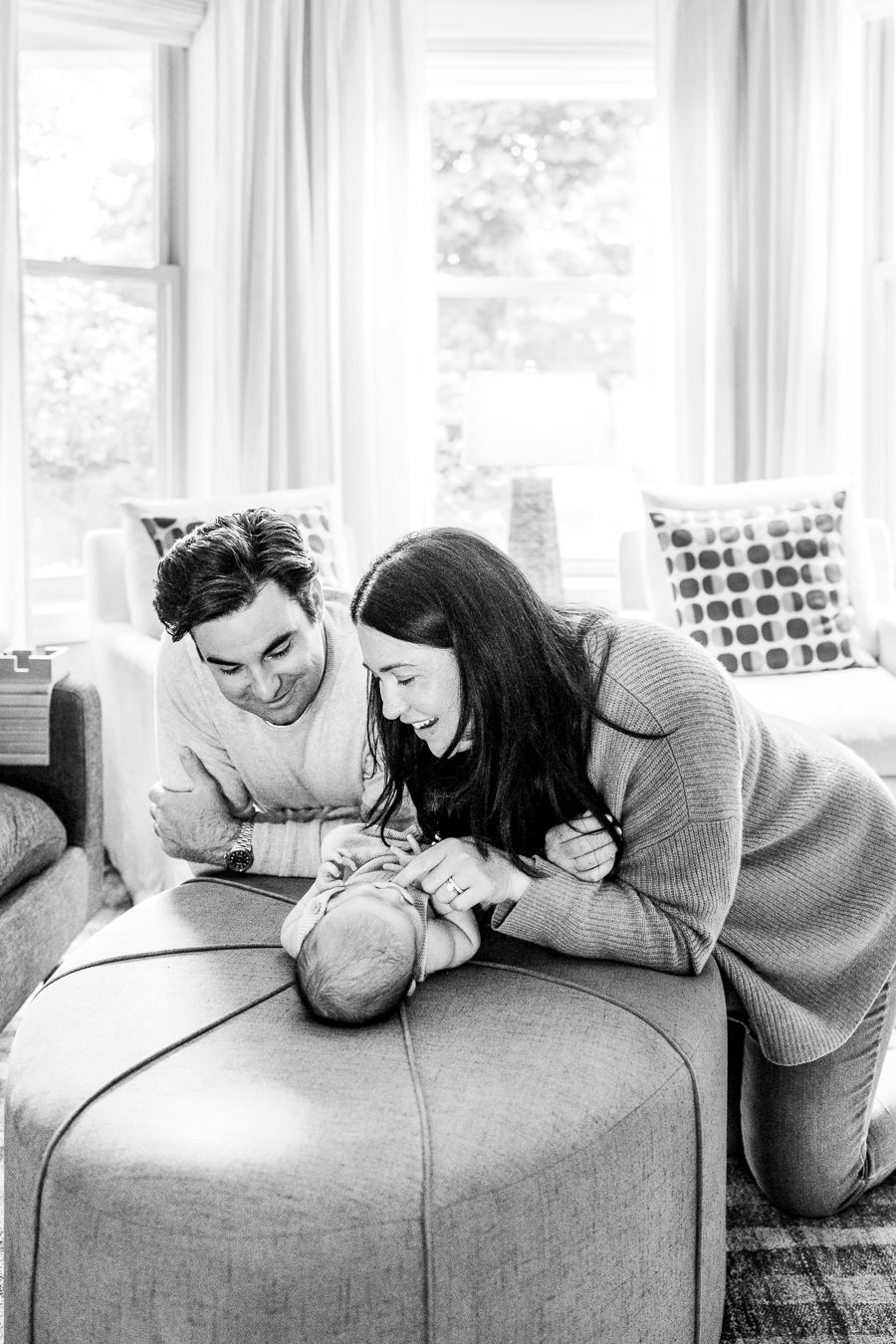 Black and white photo of parents lovingly gazing at their newborn baby on an ottoman in a bright living room.