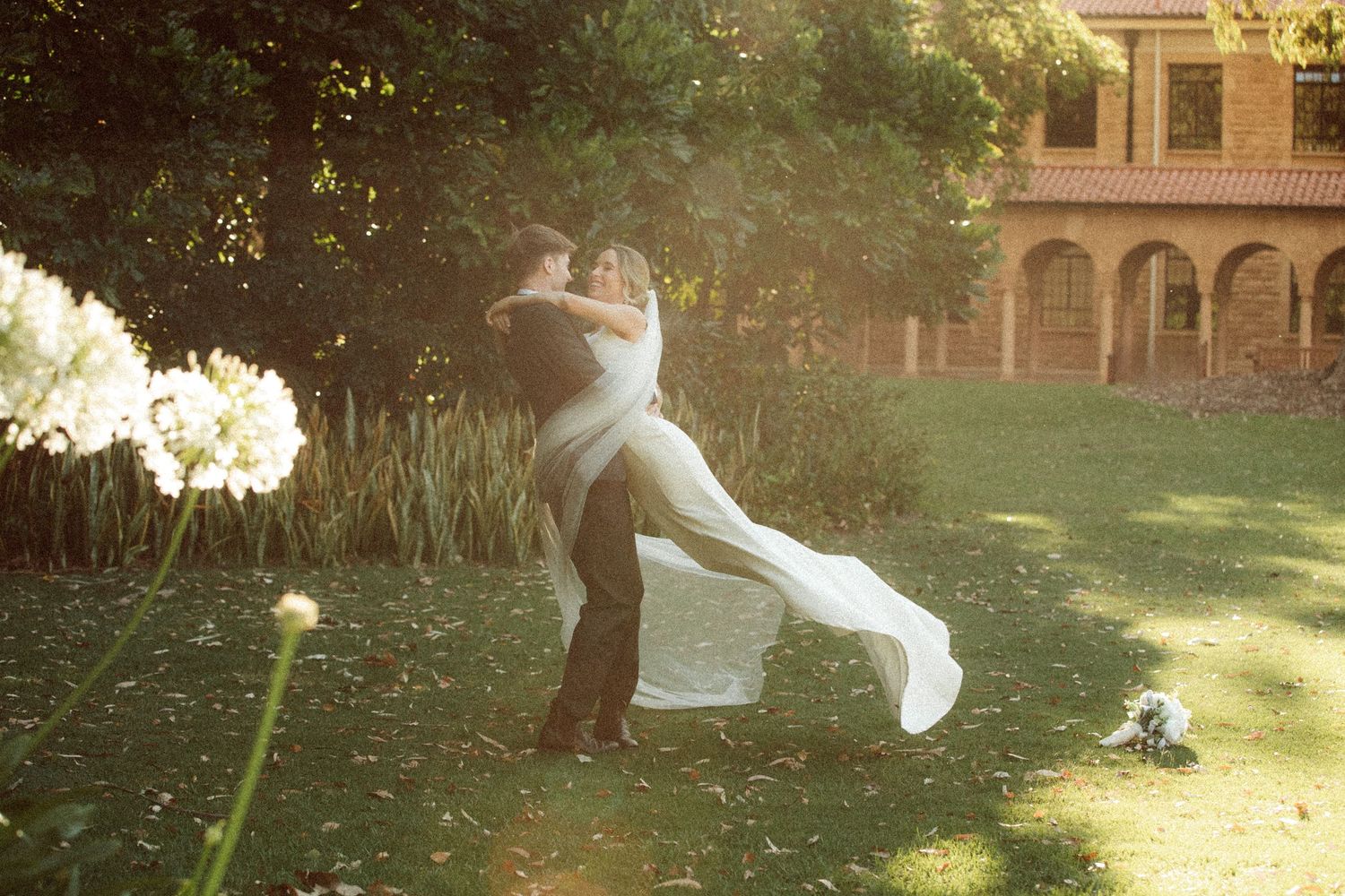 A groom lifts his bride in a sunlit garden, her white dress flowing, near a historic building.