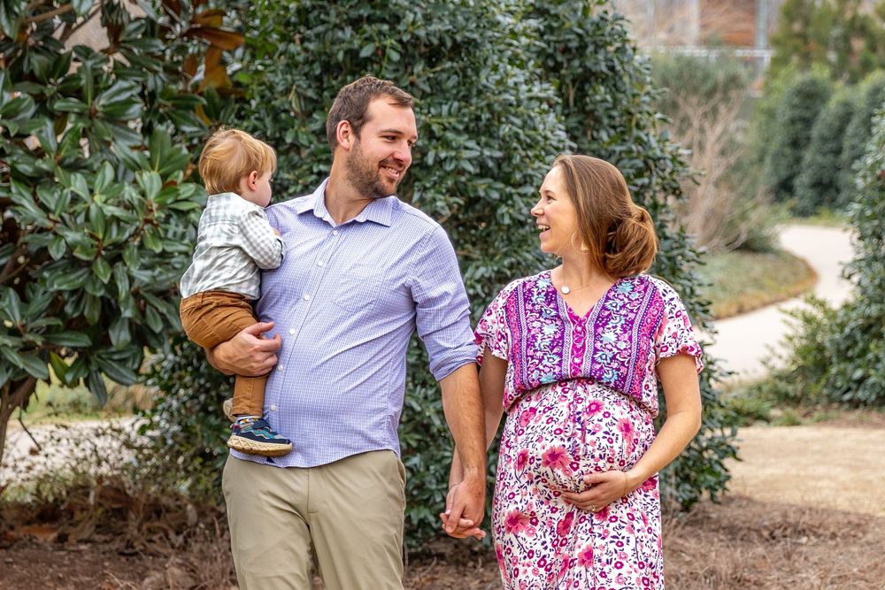 Expecting couple holds toddler son while posing outdoors in garden for maternity photo session.