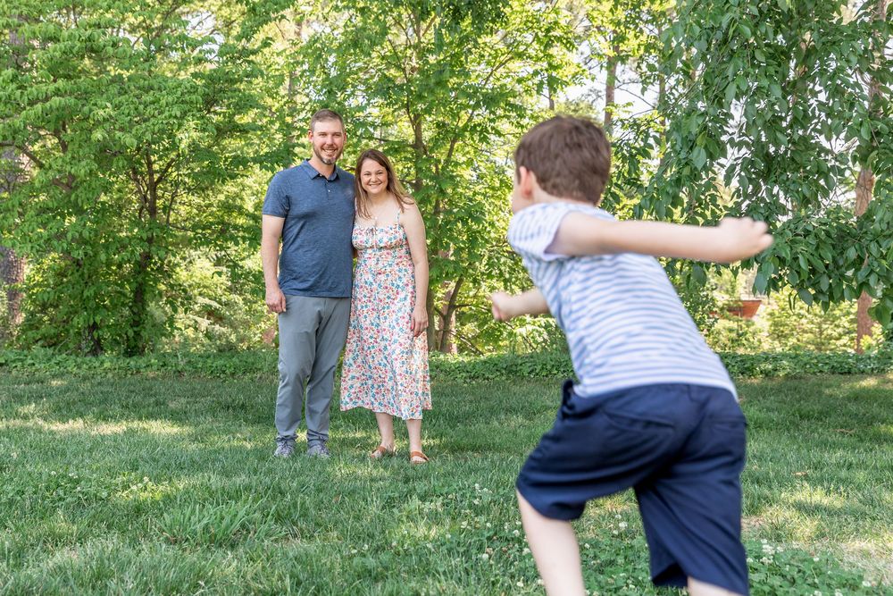 Family playing outdoors on green grass, parents smiling as child tosses frisbee in sunny backyard.