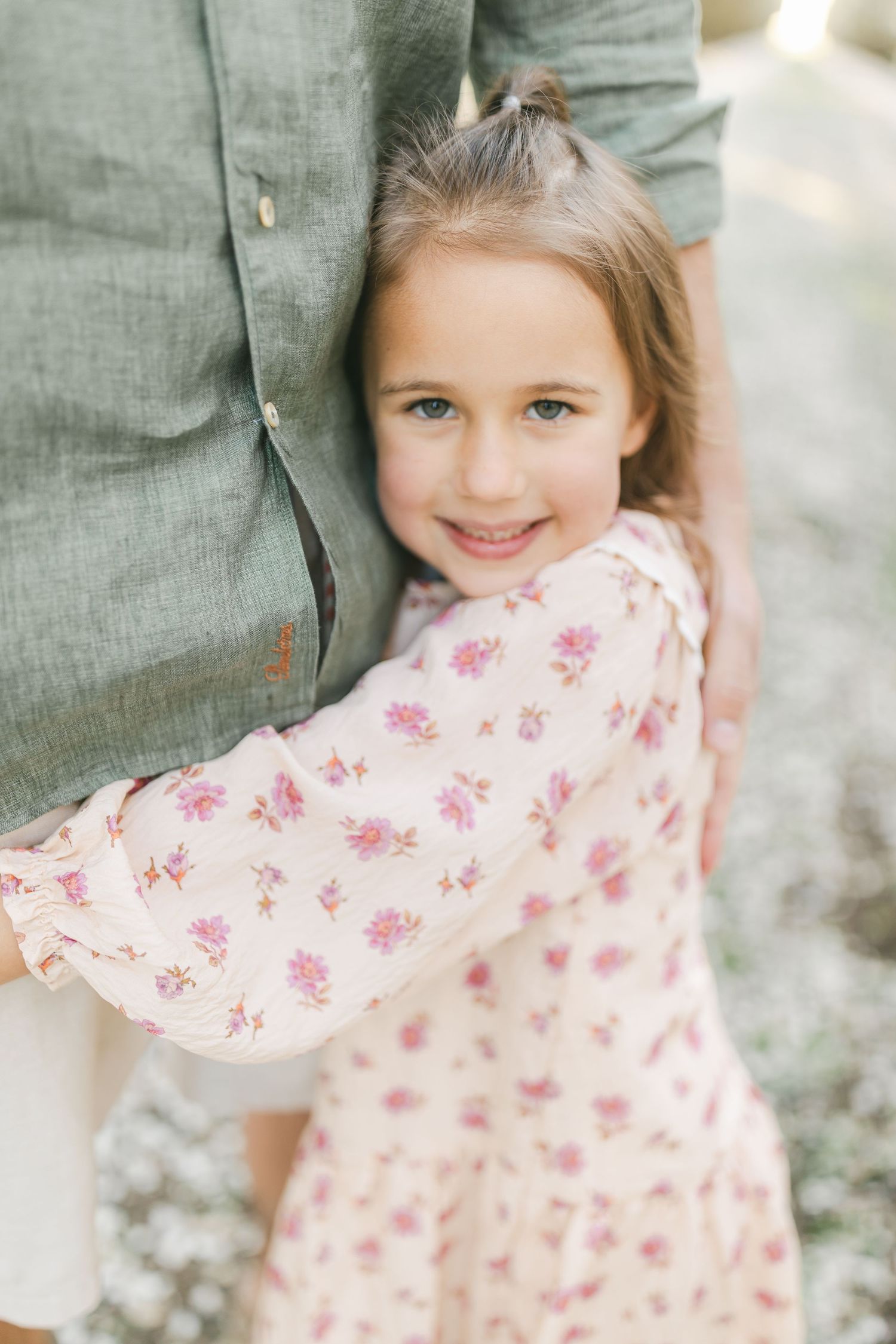 A young girl in a pink floral dress hugs an adult's leg, smiling sweetly at the camera outdoors.