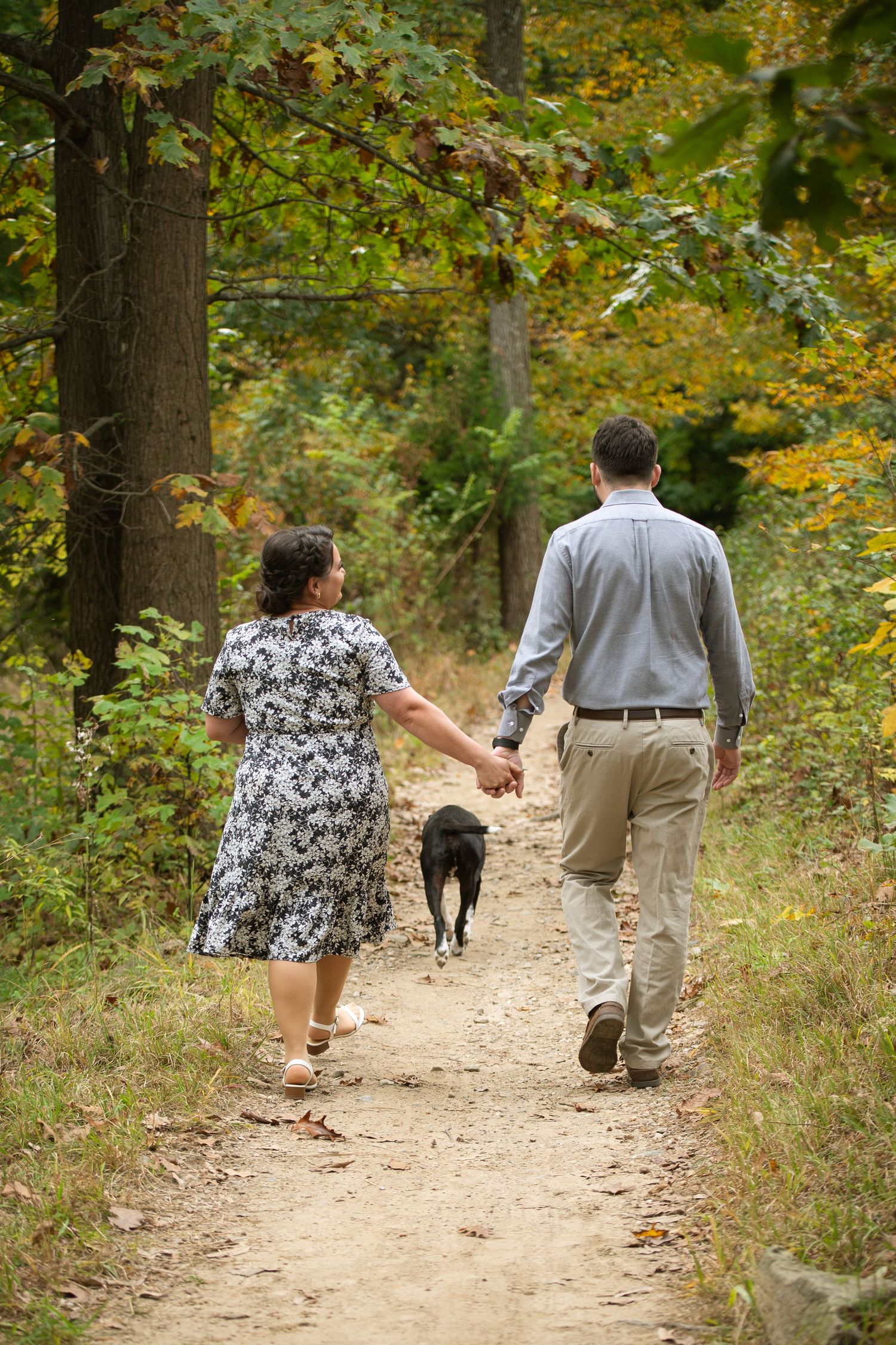 Couple holding hands walking a black dog on a forest trail surrounded by autumn foliage.