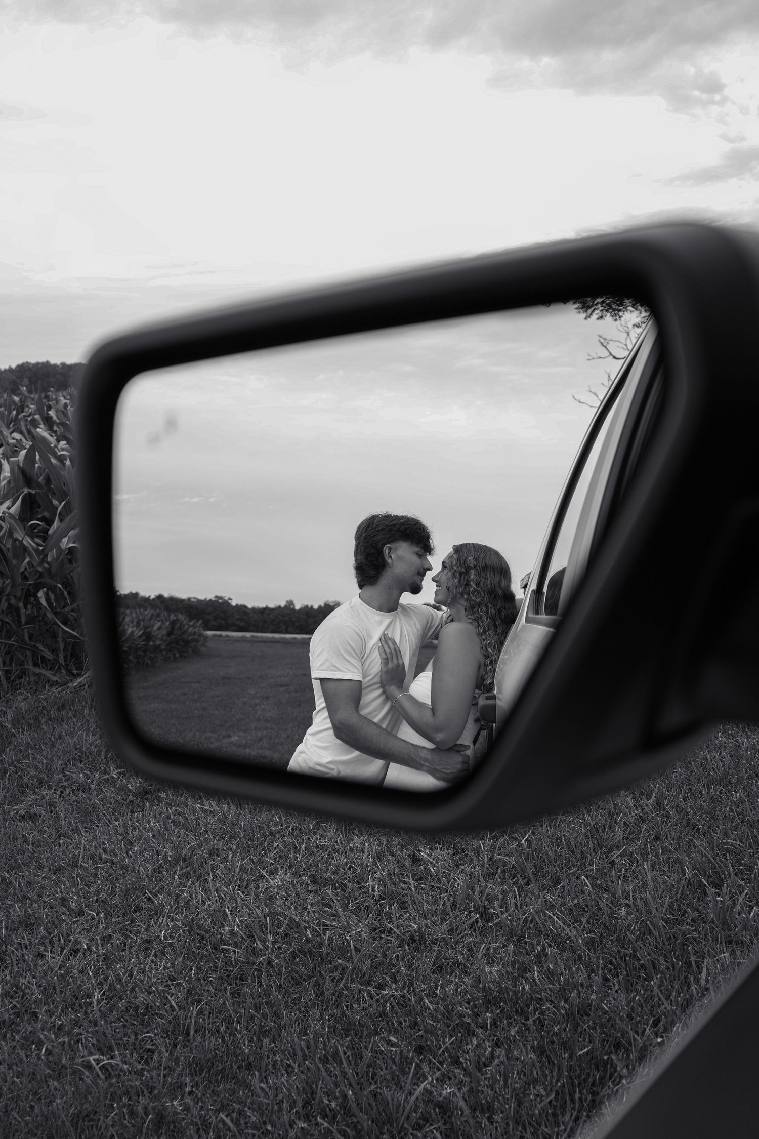 Black and white photo of a couple kissing reflected in a car side mirror with a cornfield backdrop.
