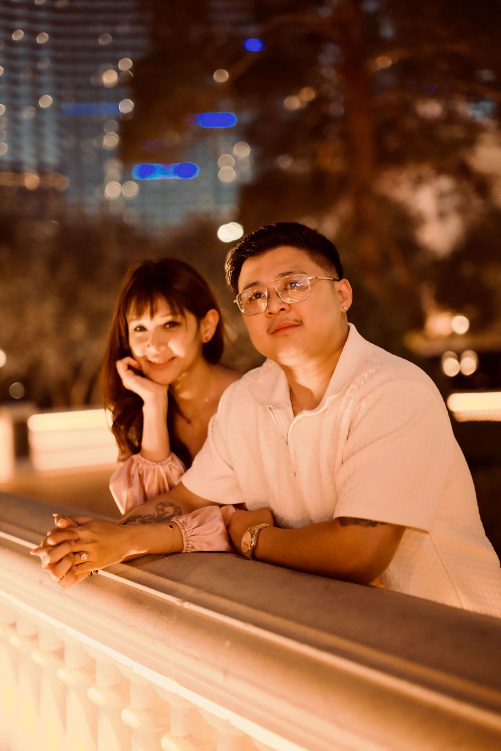 A couple leans against a railing at night with city lights bokeh in the background.