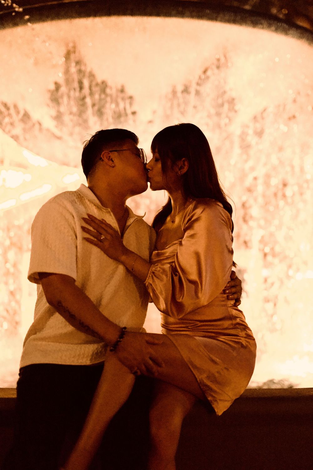 Romantic couple sharing a kiss in front of a glowing fountain at night.