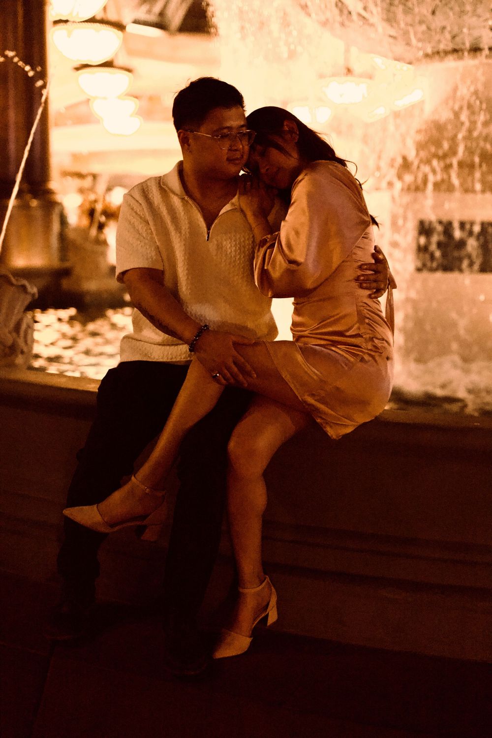 Couple sitting intimately together by a warmly lit fountain at night.