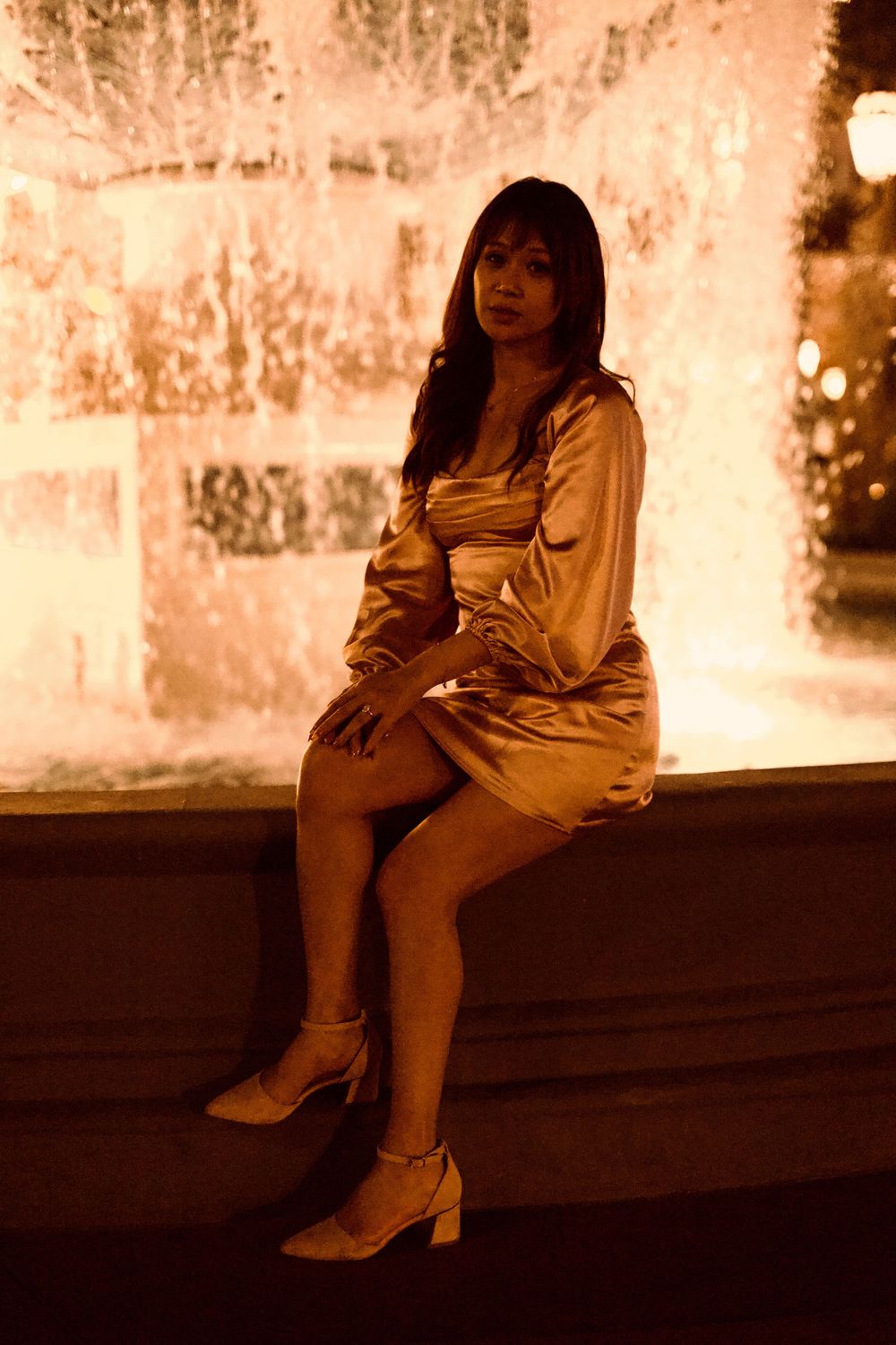 Woman in gold satin dress sitting by illuminated fountain at night in Las Vegas.