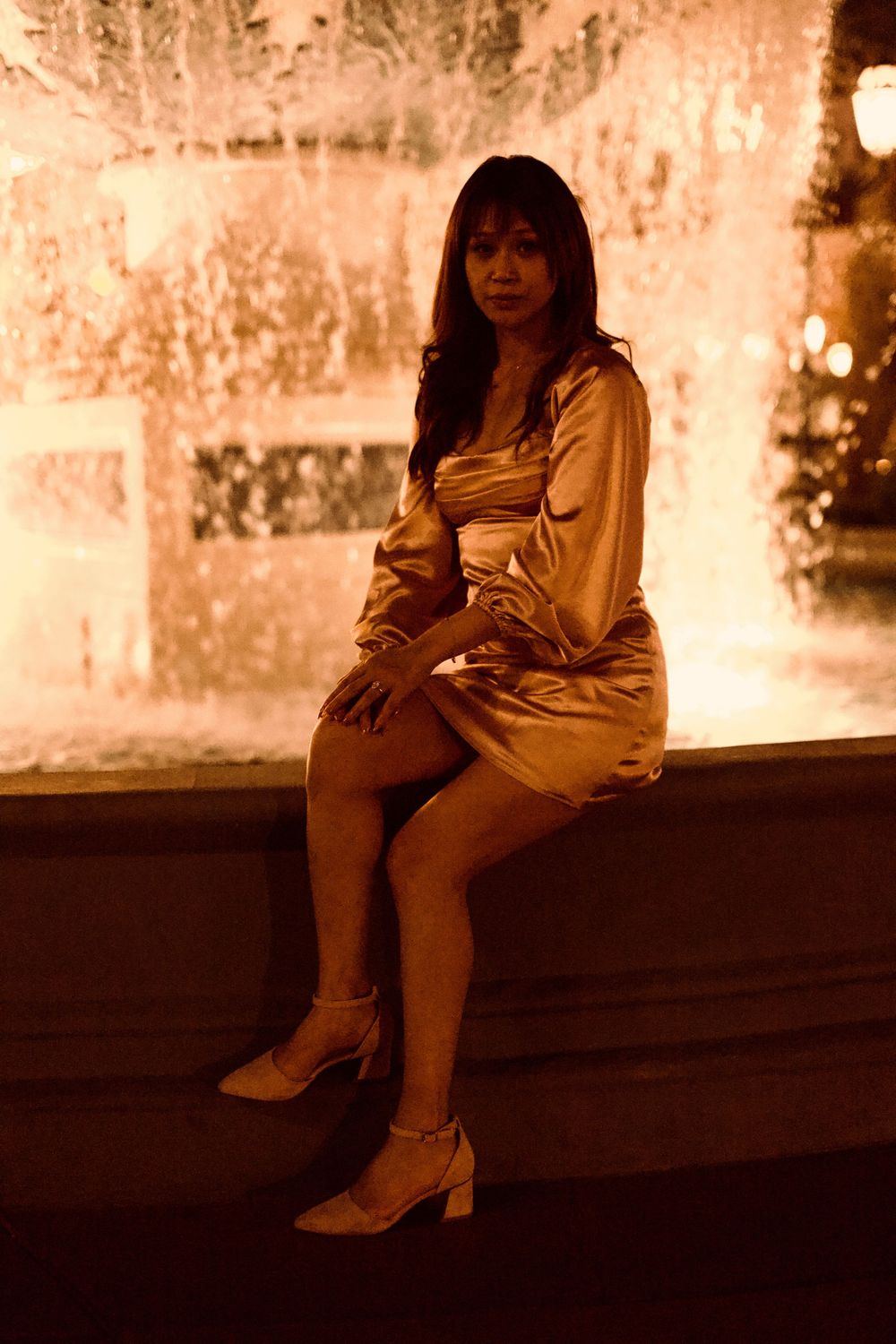 Elegant woman in gold satin mini dress sitting by illuminated fountain at night.