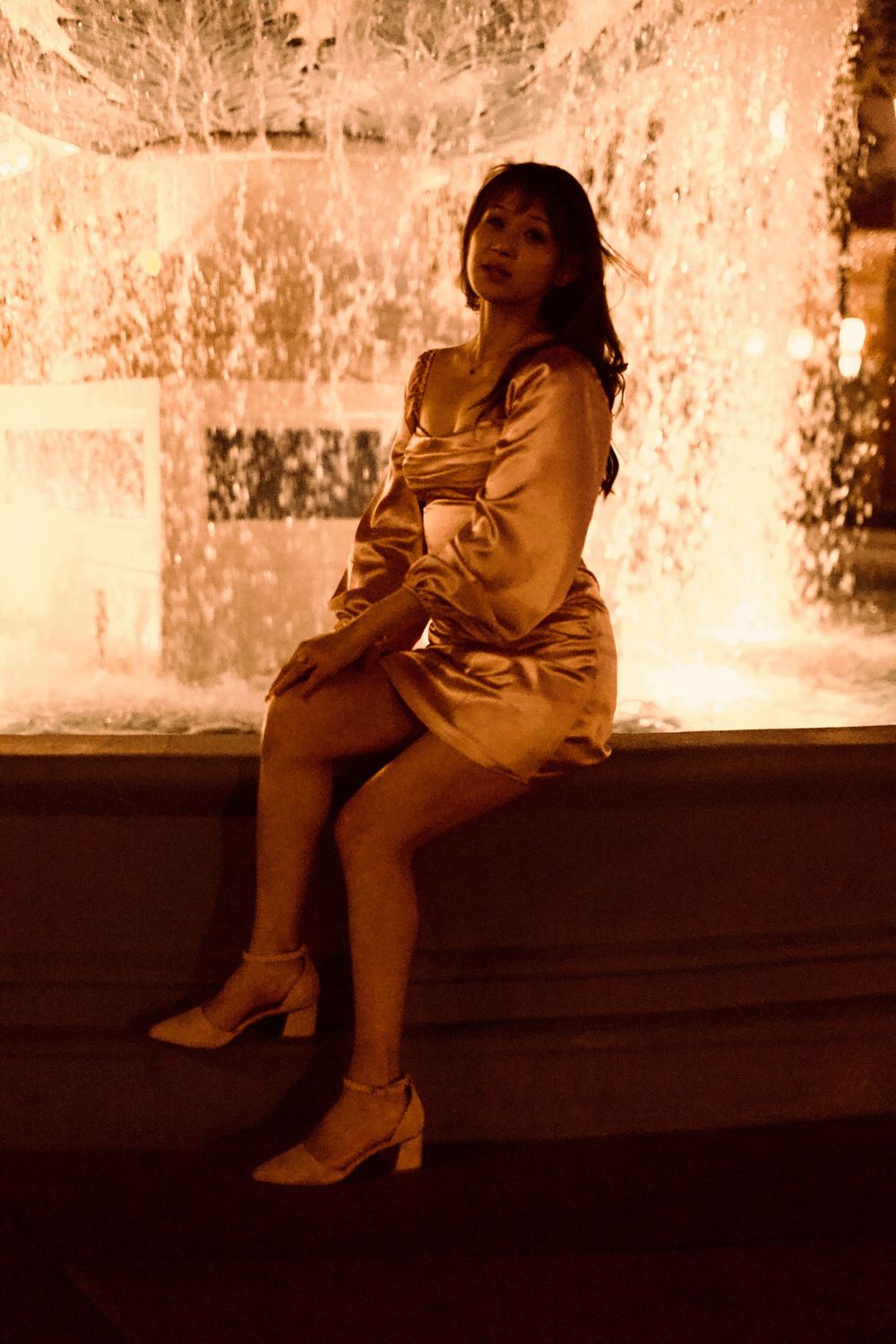 Stylish woman in golden satin dress posing by a glowing fountain at night.