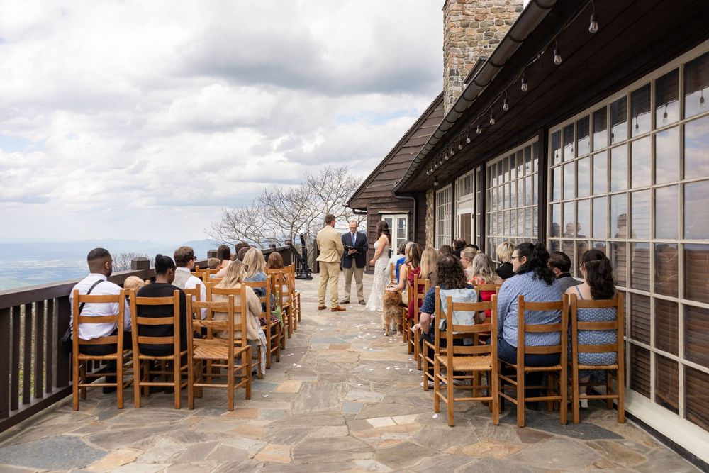 Outdoor wedding ceremony with guests seated in white chairs on a brick patio surrounded by lush green trees.