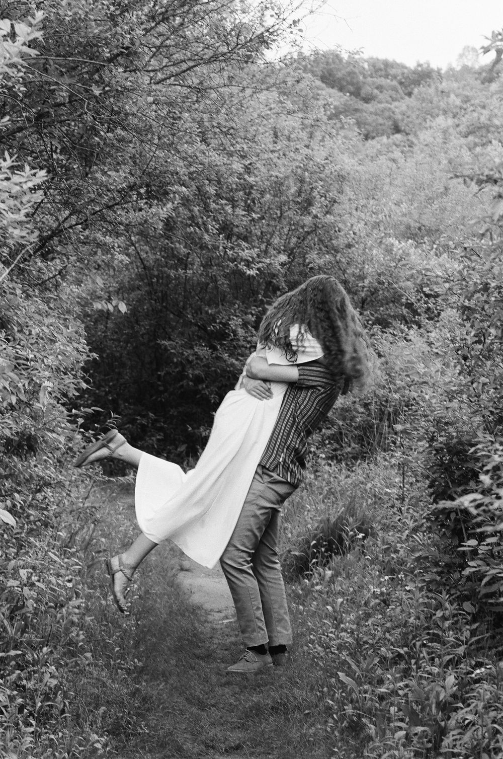 Black and white photo of a man lifting a woman in a white dress on a lush forest path.