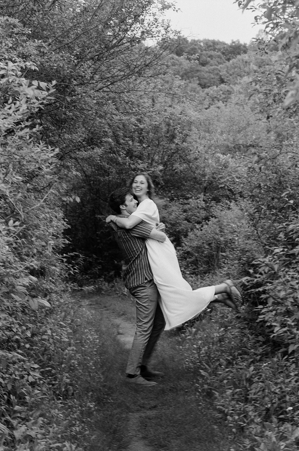 A groom lifts his smiling bride on a forest path surrounded by lush trees in a black and white wedding photo.
