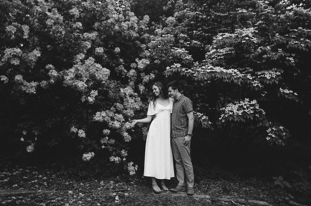 Black and white photo of a couple standing together in front of lush flowering bushes in a garden setting.