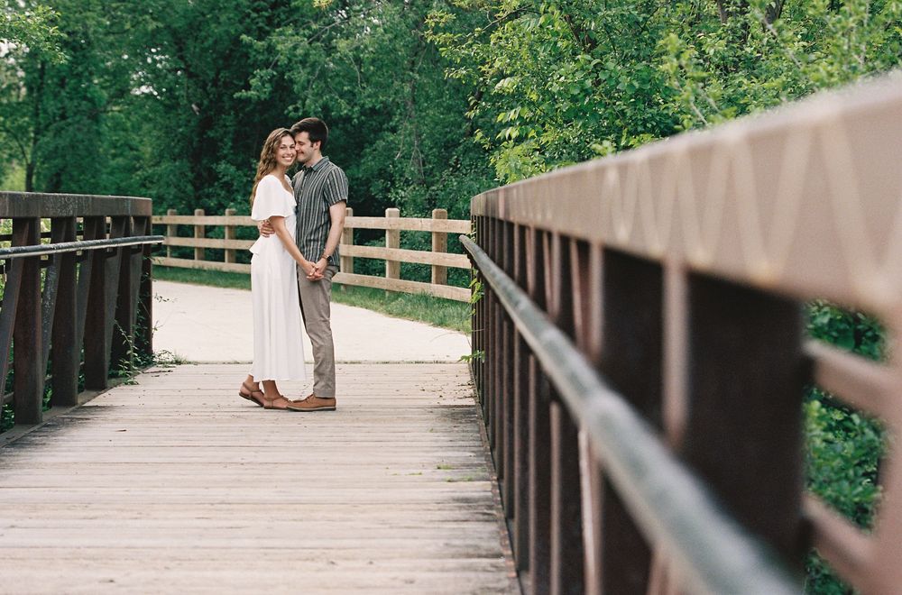 Couple sharing a romantic kiss on a wooden bridge surrounded by lush green trees.