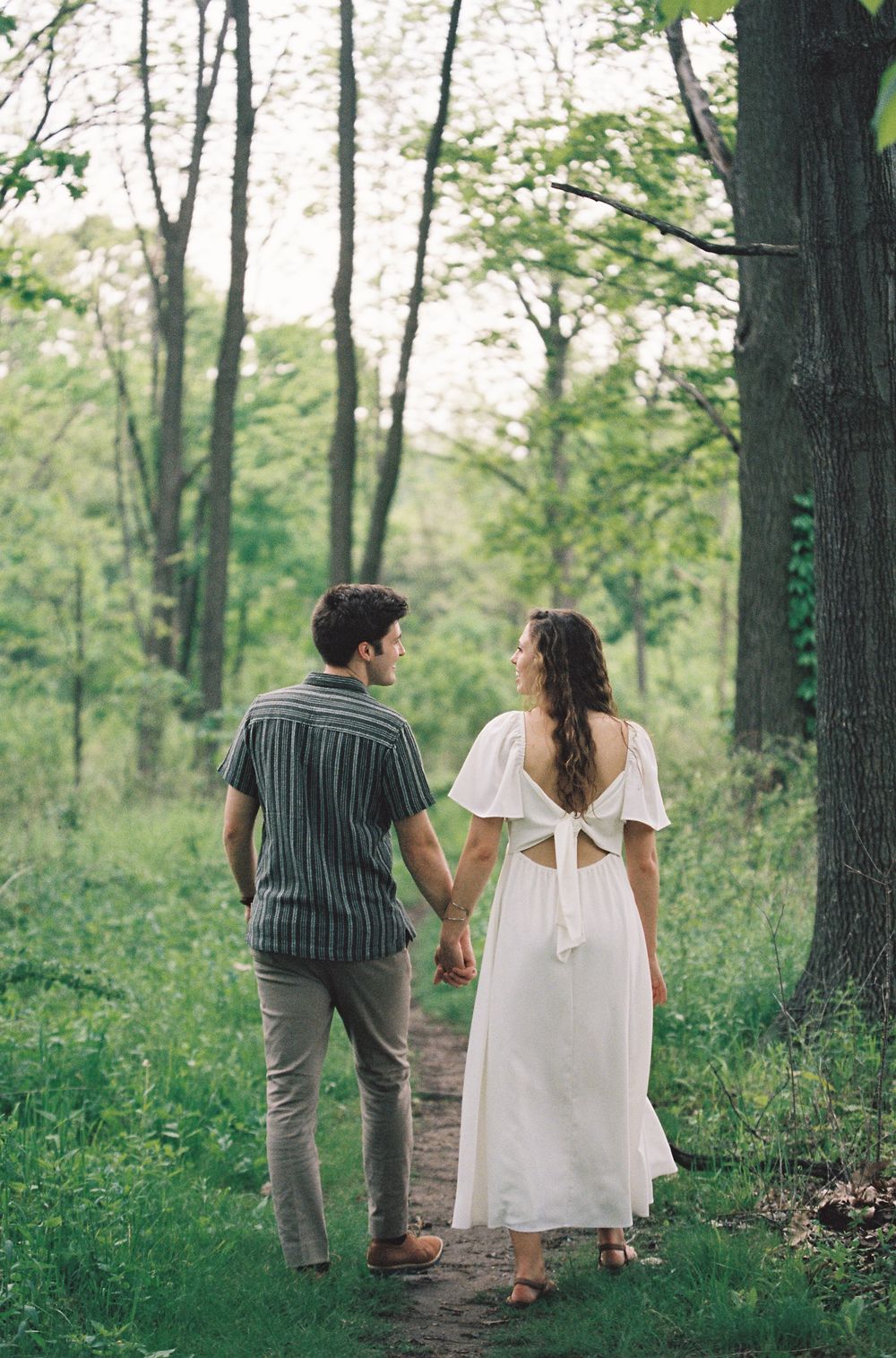 A couple holds hands walking along a lush forest path, the woman in a white dress with a bow back, the man in a striped shirt.
