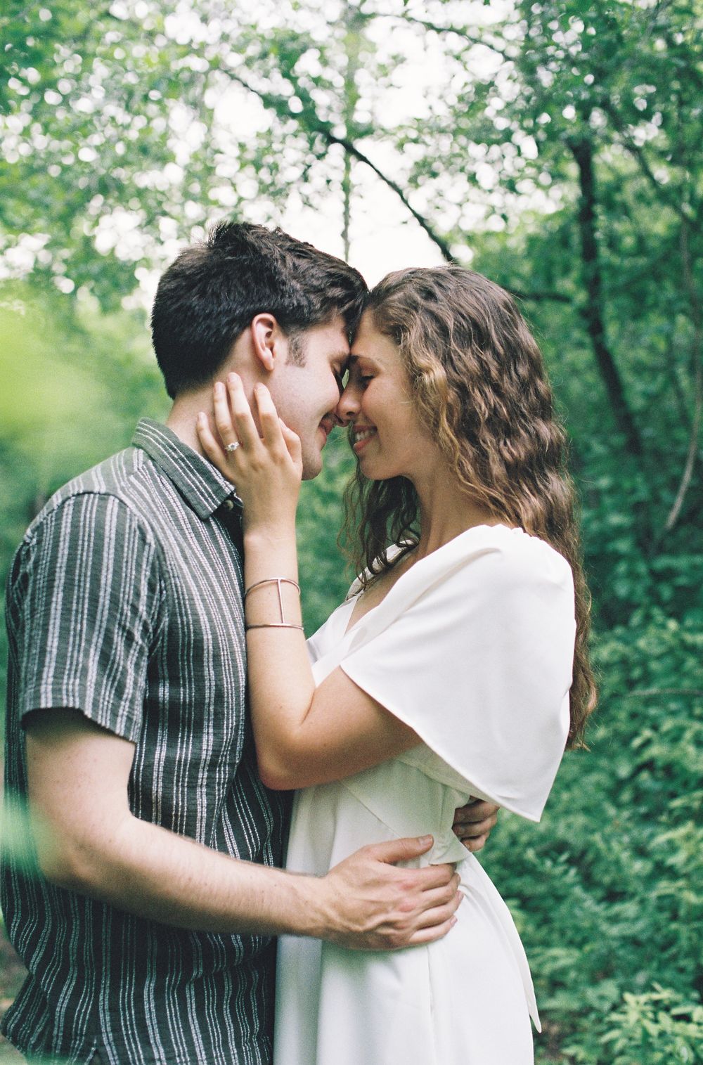 A couple shares a romantic almost-kiss moment surrounded by green trees outdoors.
