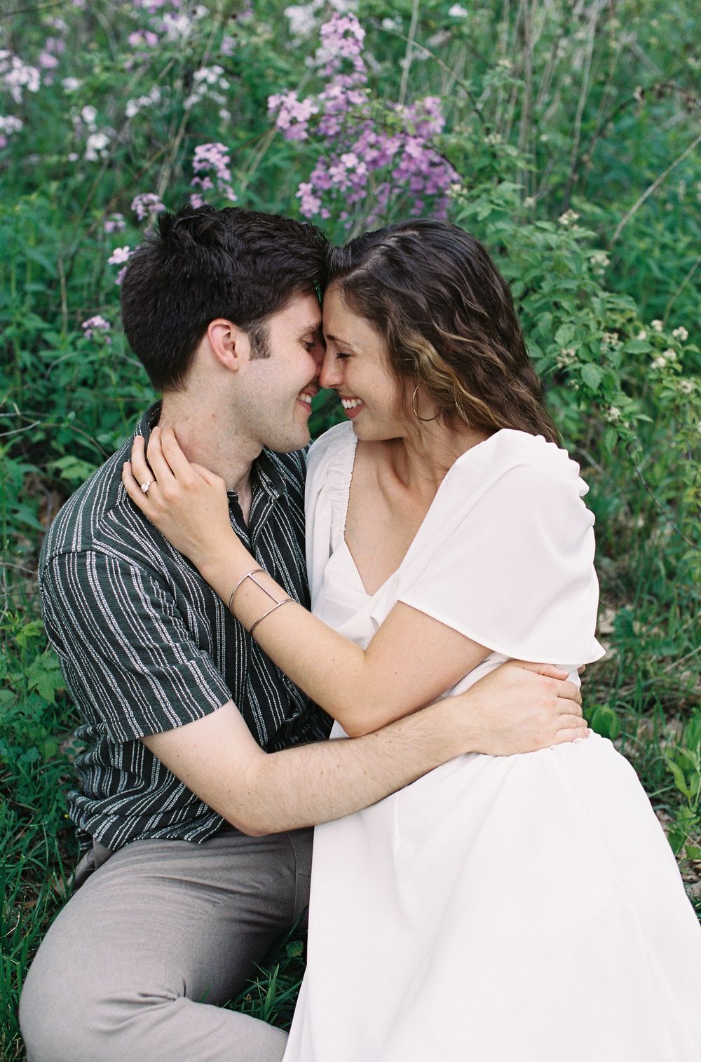 A smiling couple sits closely together in a field of wildflowers, touching foreheads tenderly.