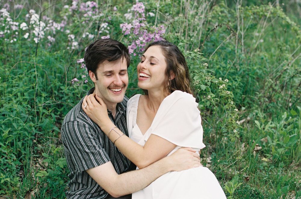 A happy couple embraces and laughs together while sitting in a field of wildflowers during an engagement session.