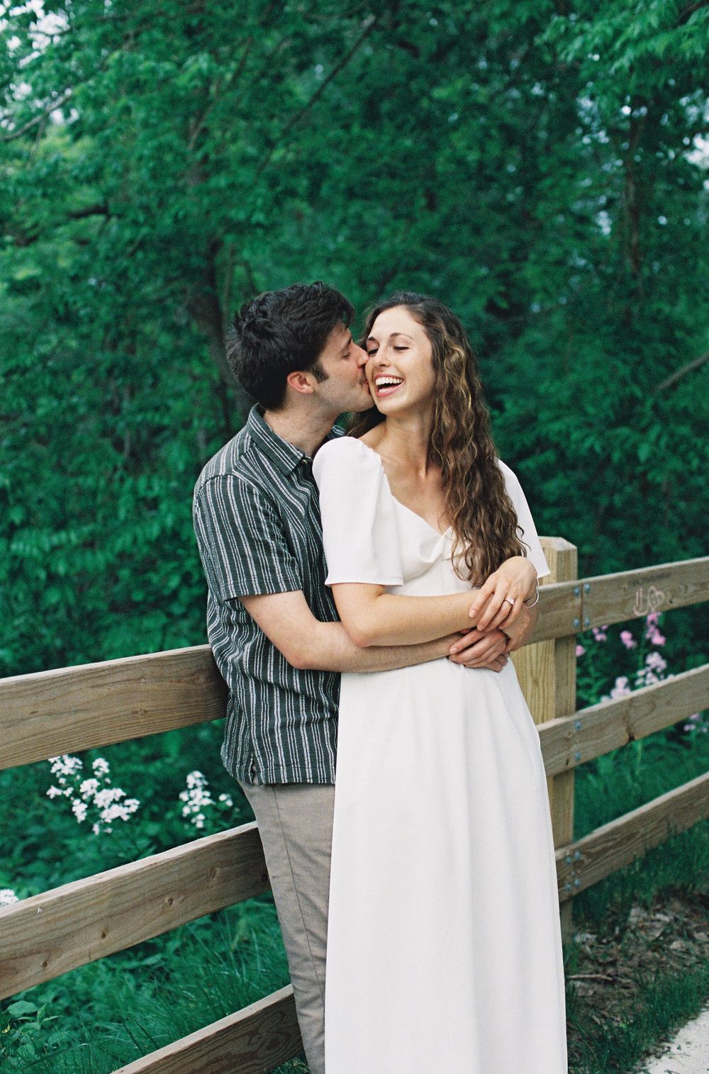 Smiling couple embracing by wooden fence surrounded by green trees, woman in white maxi dress.