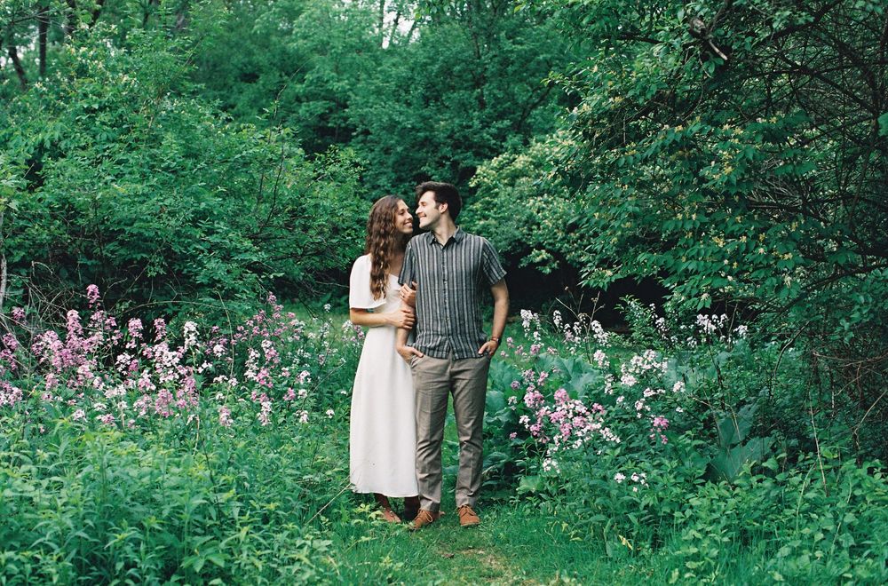 Couple standing among pink wildflowers and lush green trees in a romantic outdoor garden setting.