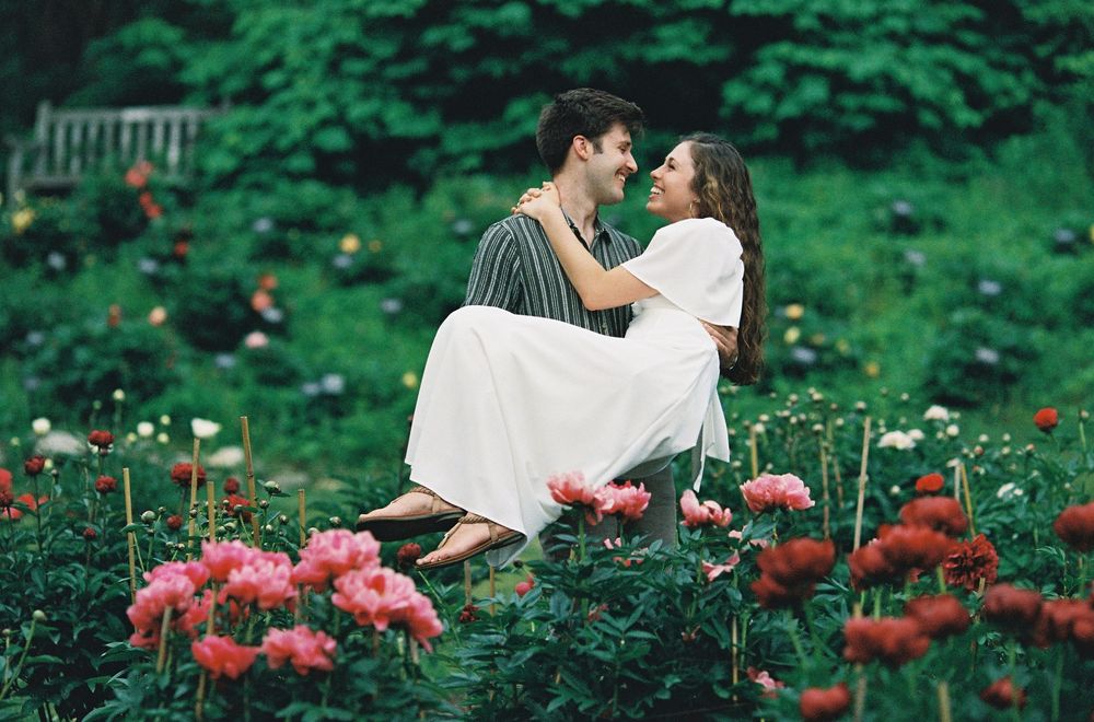A romantic couple sits together among vibrant pink peony flowers in a garden setting.