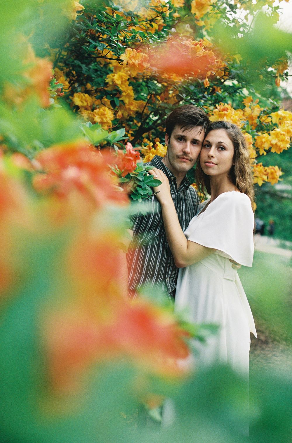 Romantic couple posing among vibrant orange and yellow flowering bushes in a garden setting.