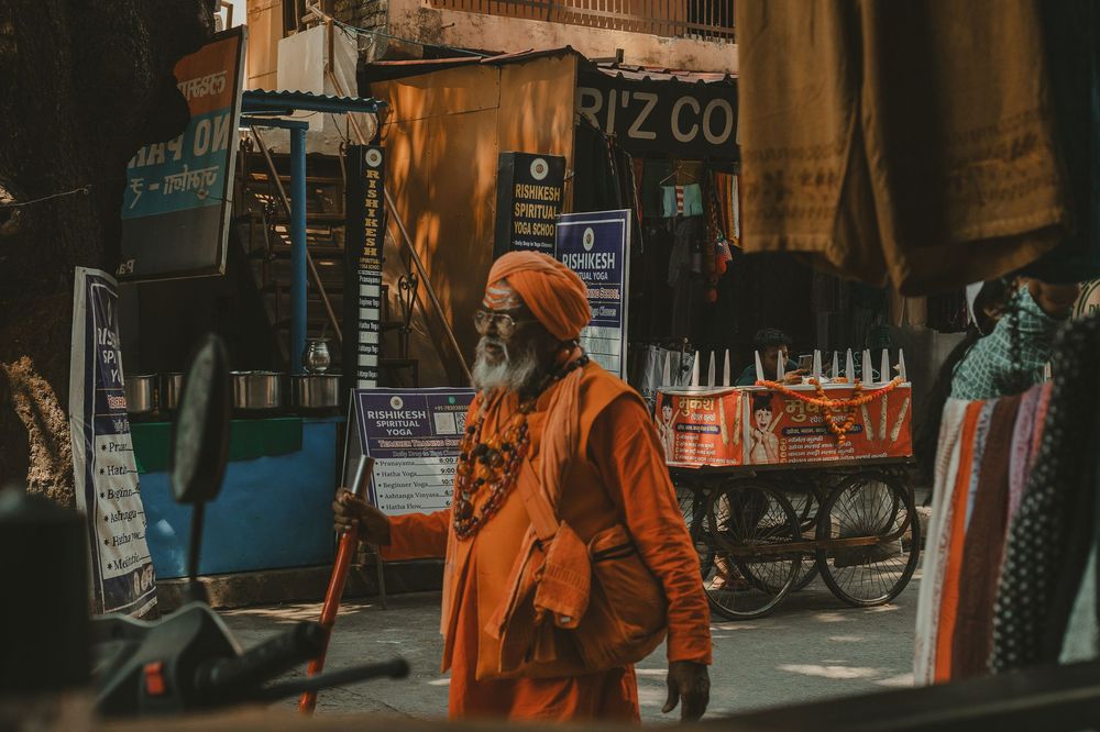 Elderly sadhu in orange robes holding a staff in a busy Indian market street.