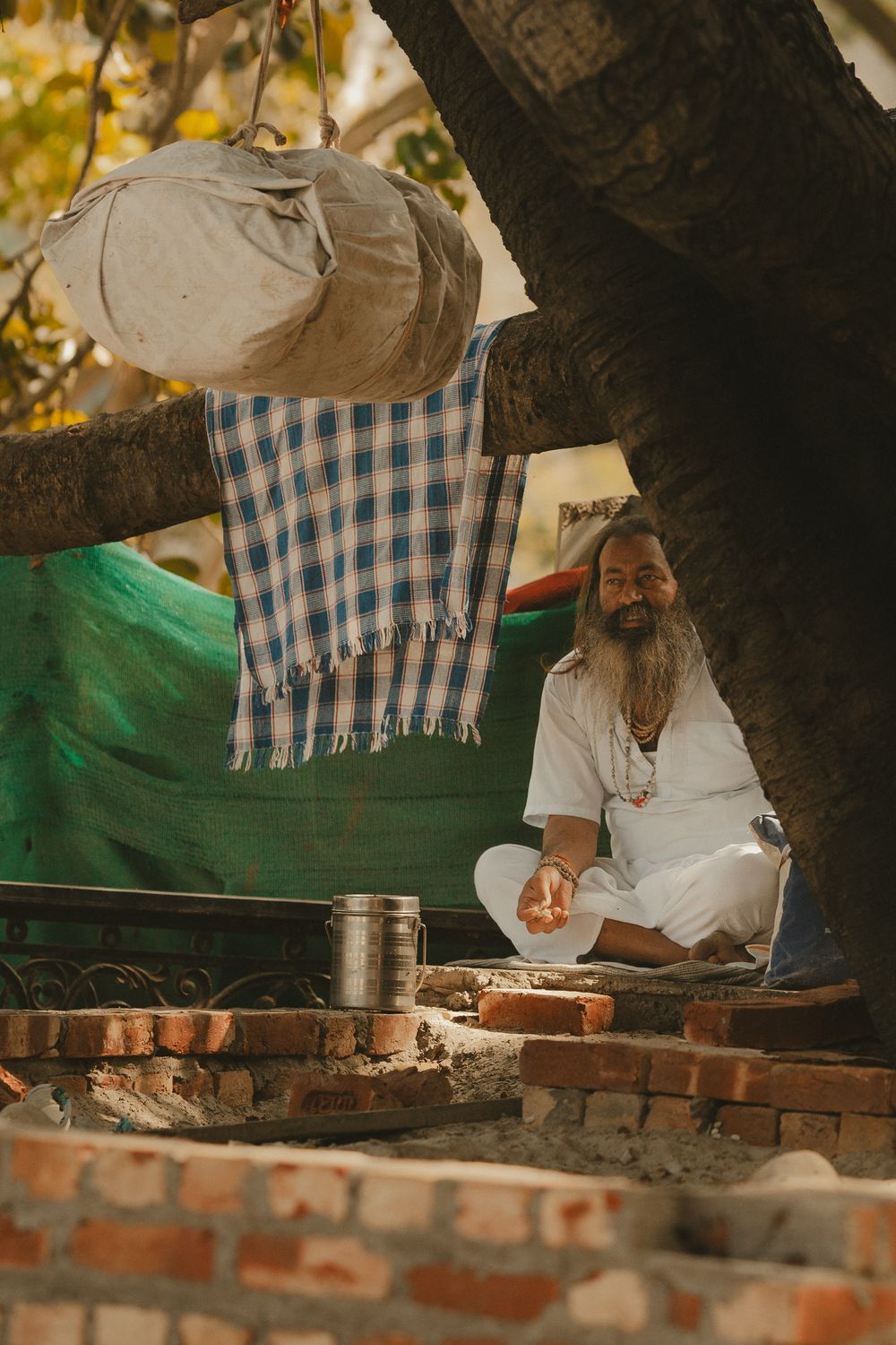 A serene Indian sadhu in white robes meditates under a tree with belongings.