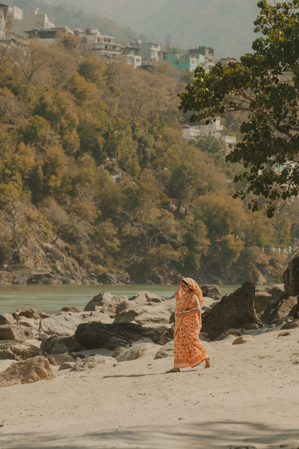 Woman in orange sari walking along a rocky beach on the Ganges River in Rishikesh, India.