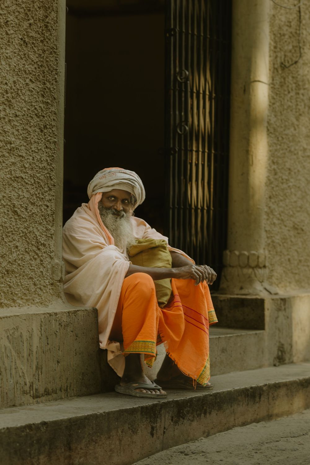 An elderly Indian sadhu in orange robes and white turban rests on temple steps.