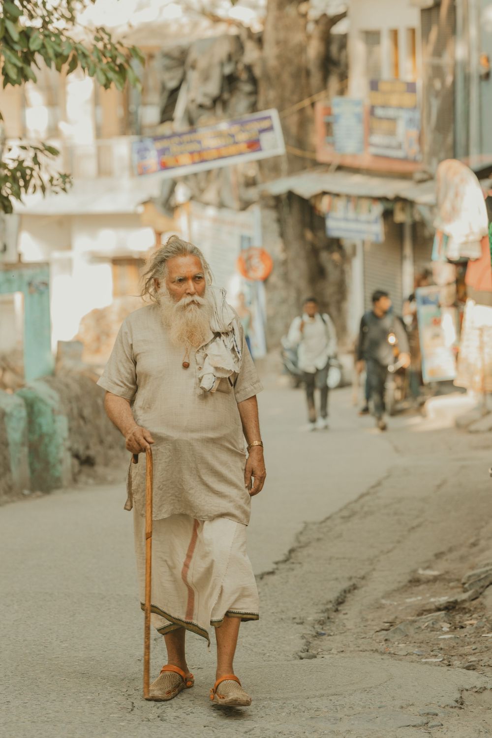 An elderly bearded Indian man walks with a wooden staff along a busy street.