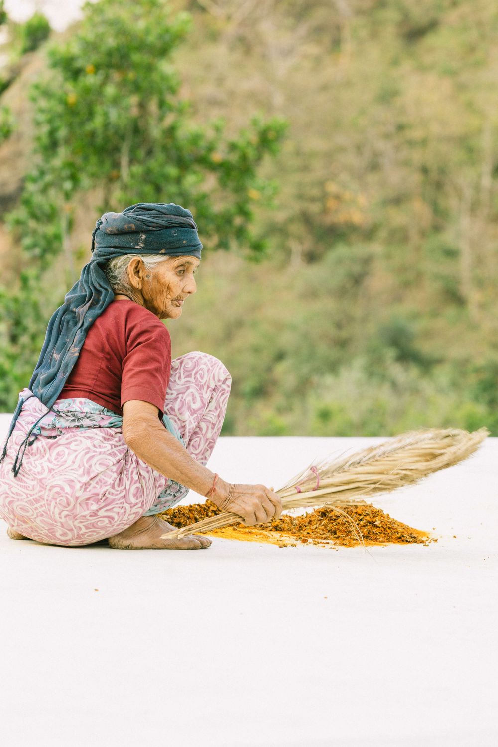 Elderly Indian woman sitting outdoors threshing grain with a broom on a rooftop.